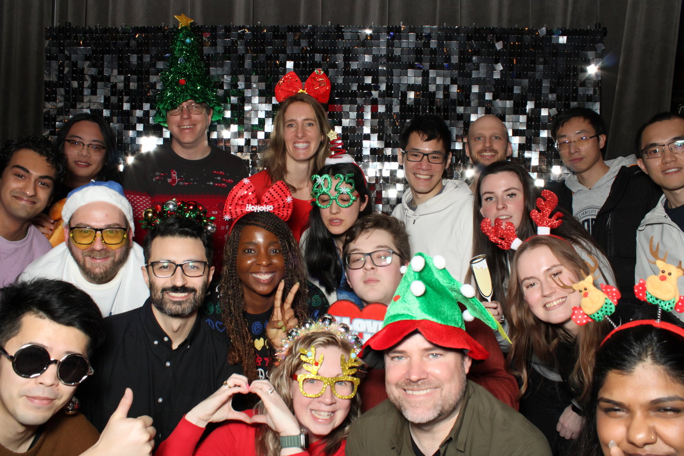 A big crowd in Christmas tree hats and festive glasses on a black sparkle wall at the ASE and SWE Holiday Lunch